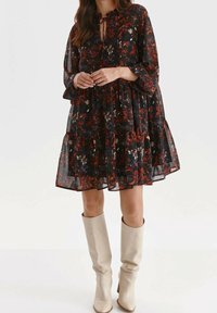 Svart blommig klänning med flödande silhuett, med rundad halsringning och långa ärmar. Accenterad med ett repknut och bärs med knähöga beiga stövlar.