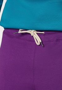 Der türkisfarbene Pullover und die lila Hose verfügen über eine Taillenpartie mit Kordelzug in Beige und silbernen Endkappen, die eine glatte, dehnbare Stoffstruktur betont.