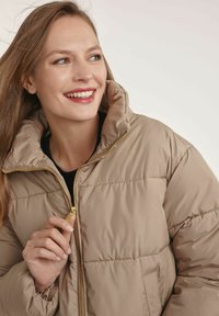 Beżowa kurtka puchowa z wysokim kołnierzem i złotym zamkiem błyskawicznym. Teksturowany materiał z wypukłymi sekcjami; model trzyma zamek.