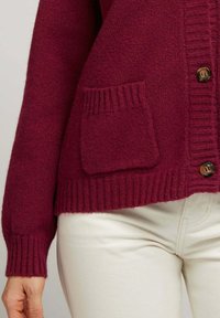 Cardigan en laine bordeaux avec une texture côtelée, doté d'une fermeture à un bouton et d'une poche plaquée sur le côté gauche.