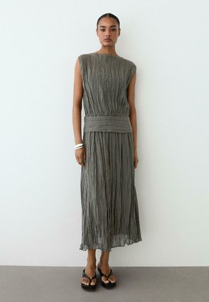 Femme portant une robe midi grise froissée sans manches, ceinture large, bracelets argentés et sandales noires à plateforme, debout contre un mur blanc.