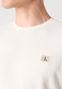Gros plan d'une personne portant une chemise blanche texturée à manches longues avec un petit écusson beige du logo Calvin Klein sur la poitrine.
