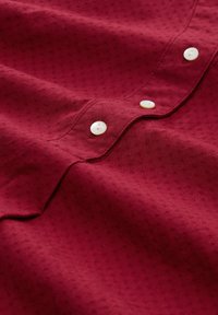 Roter texturierter Stoff mit drei weißen Knöpfen entlang einer Naht, die leicht gefaltet ist und Nähte zeigt.