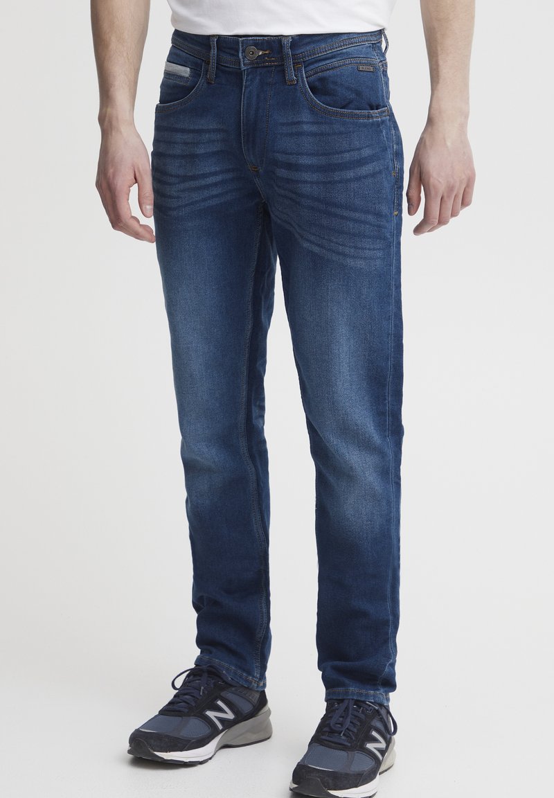 Jeans de mezclilla azul para hombre con un ajuste slim, patrón sutil de desgaste, cinco bolsillos y cierre de botón clásico. Combinados con zapatillas deportivas oscuras.