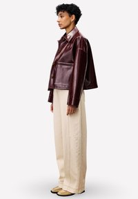 Persoon in een glanzende bordeauxrode leren jack, losse crème wijde broek en beige schoenen, staand in profiel op een witte achtergrond.