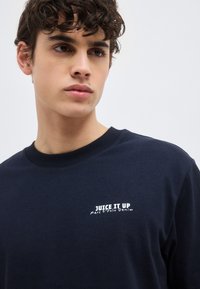 Νέος άντρας με σκούρα σγουρά μαλλιά που φοράει μπλε σκούρο φούτερ με την επιγραφή «JUICE IT UP Marc O’Polo Denim» στο στήθος.