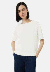 Weißes Kurzarmshirt mit weitem Ausschnitt, kombiniert mit dunkelblauen, geraden Jeans. Der Stoff wirkt glatt und leicht.