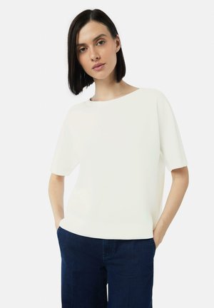 Weißes Kurzarmshirt mit weitem Ausschnitt, kombiniert mit dunkelblauen, geraden Jeans. Der Stoff wirkt glatt und leicht.