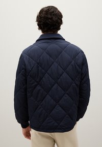 Uomo con capelli ricci scuri che indossa una giacca trapuntata blu marina e pantaloni beige, in piedi di fronte a uno sfondo chiaro uniforme.