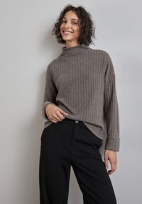 Brauner, gerippter Strickpullover mit hohem Kragen und langen Ärmeln, kombiniert mit schwarzen Hosen, vor einem grauen Hintergrund.