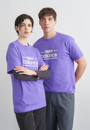 Doi tineri adulți purtând tricouri mov asortate cu inscripția „New Balance” stau sprijiniți de un fundal simplu, deschis la culoare, privind înainte cu expresii neutre.