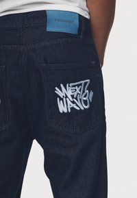Dark denim jeans with embroidered "NEXT WAVE" text in white. Features a blue PEGADOR label on the waistband and two back pockets.