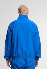 Blue lightweight jacket with a stand collar, elastic cuffs, and waistband. Textured fabric, loose fit, and simple design with no visible fasteners.