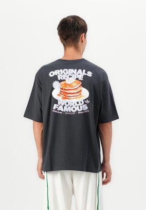 Osoba ubrana w czarną koszulkę z nadrukiem stosu naleśników oraz napisem „Originals Recipe World Famous” na plecach, w połączeniu z białymi spodniami z zielonymi paskami.