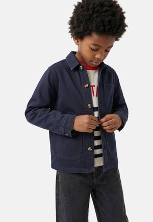 Garçon aux cheveux bouclés boutonnant une veste bleu marine, portant un pull rayé et un jean noir, debout devant un fond clair uni.