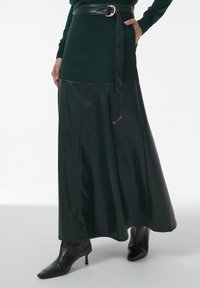 Donkergroene rok met een aansluitende top, leren accenten en een bijpassende riem. Het ontwerp loopt uit in een lange, zwierige onderkant. Zwarte hoge laarzen.