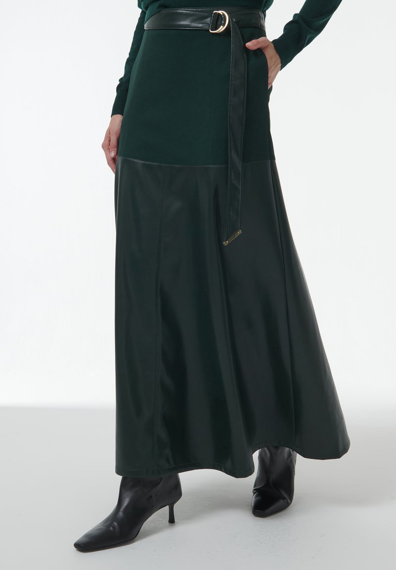 Donkergroene rok met een aansluitende top, leren accenten en een bijpassende riem. Het ontwerp loopt uit in een lange, zwierige onderkant. Zwarte hoge laarzen.