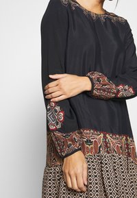 Schwarzes Kleid mit langen Ärmeln, das mit kunstvollen Ärmel- und Saumverzierungen im Paisleymuster in satten Farben versehen ist. Glatte Textur und lässige Passform.