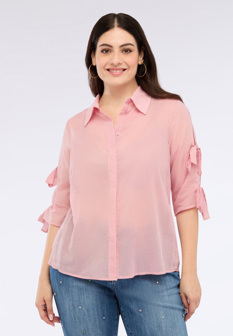 Różowa, półprzezroczysta bluzka z kołnierzykiem i zapięciem na guziki. Rękawy o długości trzy czwarte mają związane akcenty. Połączona z jasnoniebieskimi dżinsami ozdobionymi zdobieniami.