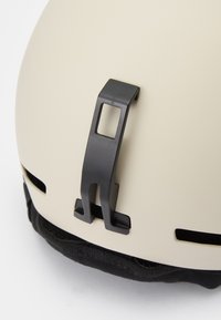 Kremasti kaciga s crnim podešavajućim isječkom i ventilacijskim otvorima, s teksturiranom crnom oblogom duž donjeg dijela za udobnost.