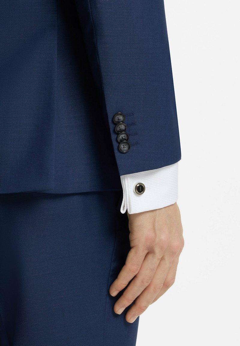 Main masculine reposant à côté d’un pantalon de costume bleu marine, manche de veste de costume avec quatre boutons noirs et poignet de chemise blanche avec un bouton de manchette rond.