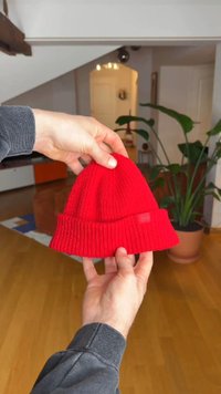 Rode gebreide muts met een geribbelde textuur en omvouwbare rand. Een klein label is voorop vastgenaaid. Vastgehouden in handen tegen een wazige interieurachtergrond.