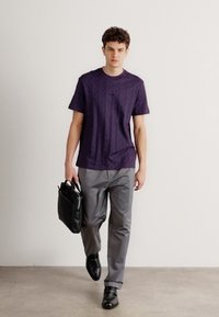 Camicetta a maniche corte viola a fantasia abbinata a pantaloni grigi e scarpe in pelle nere, mentre porta una borsa nera, camminando su uno sfondo neutro.