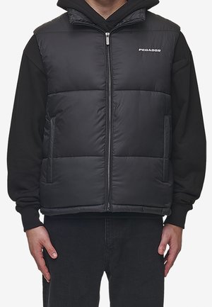 Czarna bezrękawnikowa kamizelka puchowa z przednim zamkiem i logo „PEGADOR” noszona na czarnej bluzie z kapturem i czarnych spodniach.