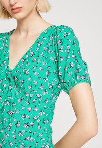 Zielona sukienka w kwiaty z krótkimi bufiastymi rękawami, z wzorem różowych i czarnych kwiatów, wiązaniem przy dekolcie i gładką fakturą.