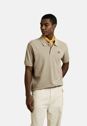 Uomo con polo beige, pantaloni bianchi e foulard giallo decorato al collo, in piedi con una mano dietro la schiena.