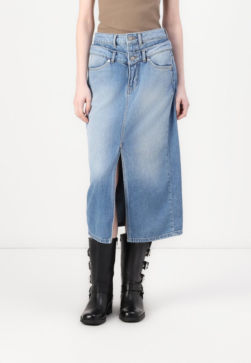 Fustă din denim de culoare albastru deschis, cu o deschidere frontală, închidere cu două nasturi și bucle pentru curea. Asortată cu cizme negre până la genunchi.