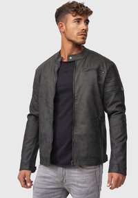 Chaqueta de cuero sintético negra con un corte ajustado, cuello alto, bolsillo con cremallera y detalles acolchados en los hombros. Llevada sobre una camisa negra.