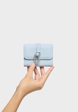 Main tenant un petit portefeuille en cuir bleu clair avec un fermoir ovale argenté sur un fond uni.