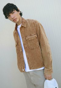 Giacca di corduroy marrone con due Tasche frontali, chiusura a bottoni e vestibilità rilassata, indossata sopra una T-shirt bianca e pantaloni grigio chiaro.