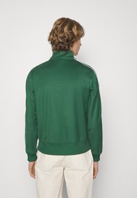 Veste zippée verte avec un col haut, ornée de bandes latérales blanches et d'un ourlet côtelé. Tissu doux avec une texture lisse.
