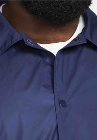 Mörkblå vindjacka med en slät struktur, försedd med tryckknappar och en klassisk kragsdesign. Vit skjorta synlig nedanför.