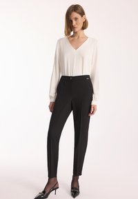 Biała bluzka z długim rękawem, z dekoltem w kształcie litery V i detalami z guzikami, zestawiona z dopasowanymi czarnymi spodniami z wyprasowanym przodem i złotym akcentem.