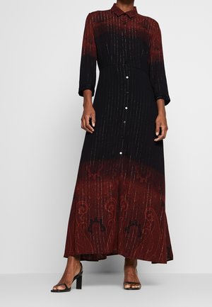 Femme portant une robe maxi à manches longues et boutonnée, avec un dégradé noir et rouge foncé et de subtiles lignes verticales scintillantes, associée à des talons noirs.