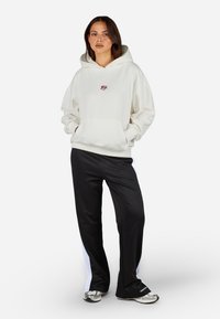Sudadera color crema con un bolsillo tipo canguro, combinada con pantalones de chándal negros que tienen franjas blancas laterales. La tela parece suave y cómoda.