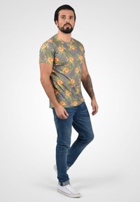 Grå t-shirt med gul og orange blomsterdesign, parret med mørkeblå jeans og hvide sneakers med røde detaljer. Glat stofoverflade.