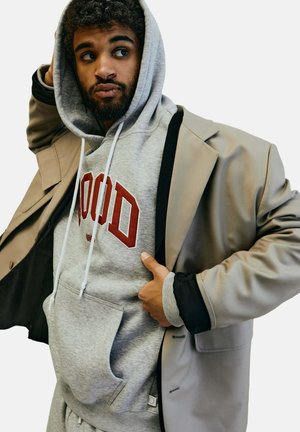 Grauer Hoodie mit roter Schrift unter einem beigen Blazer, der eine glatte Textur und eine ausgeprägte Tasche aufweist. Der Hoodie hat eine Kapuze mit Kordelzug.
