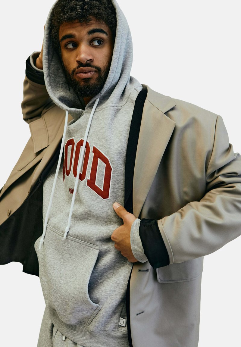 Grauer Hoodie mit roter Schrift unter einem beigen Blazer, der eine glatte Textur und eine ausgeprägte Tasche aufweist. Der Hoodie hat eine Kapuze mit Kordelzug.