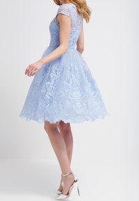 Ljusblå spetsklänning med korta ärmar, figursydd liv och utsvängd kjol. Har intrikata blommönster och en vågig nederkant. Silverfärgade klackar.