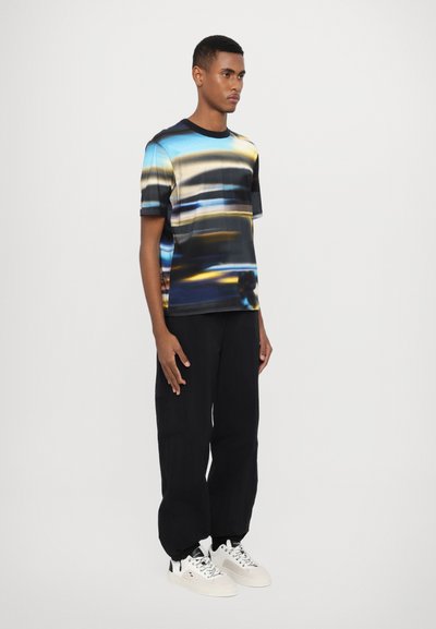 Short-sleeve t-shirt features a multicolored abstract print with blue, yellow, and black hues, made of soft fabric, paired with black pants and white sneakers.