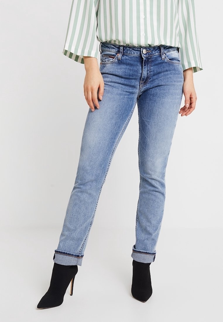 Kvinna som bär en grön och vit rutig skjorta, blå jeans och svarta klackade ankelstövlar, stående med händerna på höfterna.