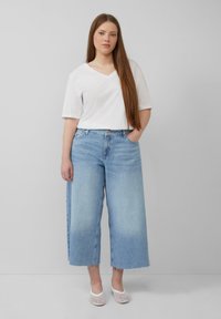 Biała koszulka z dekoltem w serek w połączeniu z jasnoniebieskimi spodniami typu wide-leg z postrzępionym dołem. Przezroczyste buty z siateczki dopełniają stylizację.