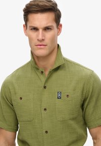 Camisa de botones de manga corta en color verde oliva con una tela texturizada, dos bolsillos en el pecho y un parche con el logo en el bolsillo izquierdo.