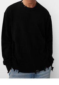 Schwarzer übergroßer Strickpullover mit fransigen Akzenten am Kragen und Saum. Rippbündchen und unterer Rand. Getragen mit hellblauen Jeans.