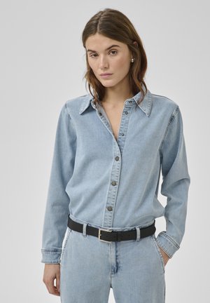 Femeie îmbrăcată într-o cămașă din denim albastru deschis și blugi cu curea neagră, stând în fața unui fundal gri simplu.