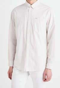 Koszula z guzikami w jasnym kremowym kolorze z cienkimi pionowymi różowymi paskami. Posiada klasyczny kołnierz, długie rękawy oraz kieszonkę na piersi.